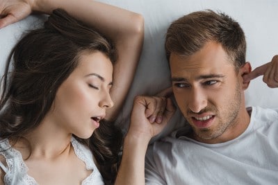man lays in bed with his fingers in his ears next to snoring wife